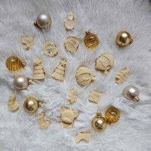 Božični dišeči voski v oblikah drevesc, Božičkov in snežakov, položeni med zlate bunkice na mehki beli podlagi – topla praznična kolekcija Christmas Mix.