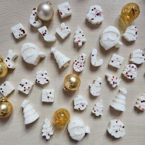 Božični dišeči voski v oblikah smrečic, snežakov in božičkov, posuti z rdečim bleščicami, poleg zlatih bunkic – praznična kolekcija Cinnamon & Apple.