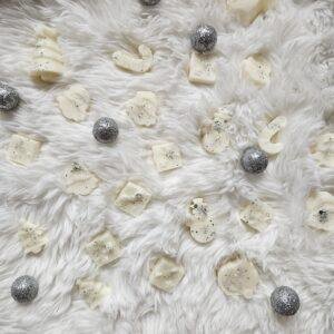 Božični dišeči voski v obliki snežakov, daril in božičnih dreves, posuti z bleščicami, položeni na belo mehko podlago s srebrnimi okraski.